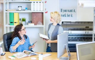 Two women converse in an office with a wide format printer in the background.