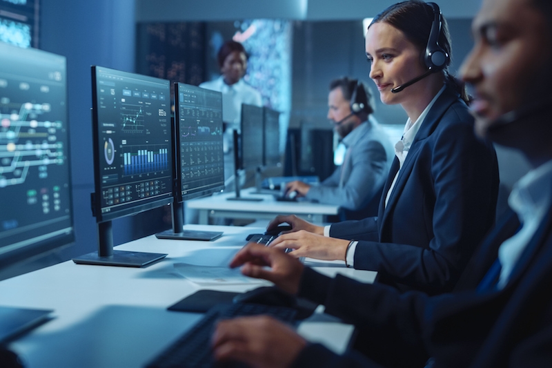 Happy Confident Technical Customer Support Specialist Having a Headset Call while Working on a Computer in a Dark Monitoring and Control Room Filled with Colleagues and Display Screens. Happy Confident Technical Customer Support Specialist Having a Headset Call while Working on a Computer in a Dark Monitoring and Control Room Filled with Colleagues and Display Screens.