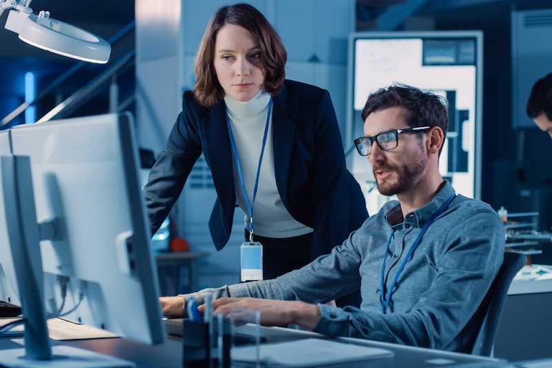 Futuristic Machine Engine Development Engineer Working on Computer at His Desk, Talks with Female Project Manager. Team of Professionals Working in the Modern Industrial Design Institution