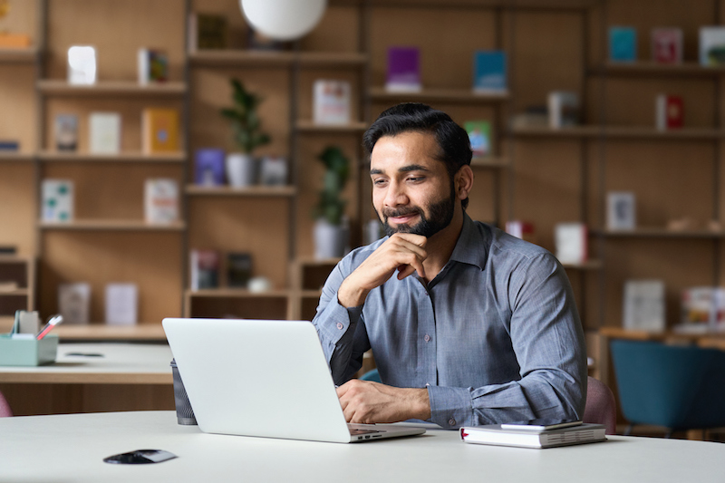 Smiling bearded indian businessman working on laptop in modern office lobby space. Young indian student using computer remote studying, watching online webinar, virtual training on video call meeting. Smiling bearded indian businessman working on laptop in modern office lobby space. Young indian student using computer remote studying, watching online webinar, virtual training on video call meeting.
