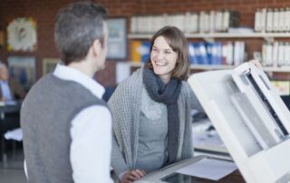 A businesswoman and businessman stand in front of one of their office copiers making a copy.