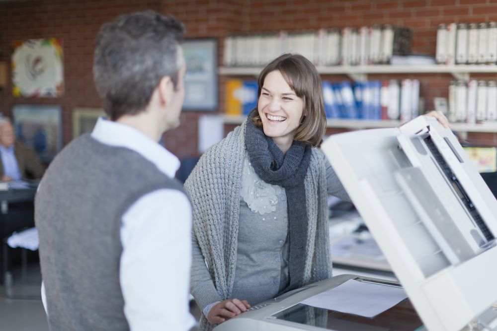 A businesswoman and businessman stand in front of one of their office copiers making a copy.