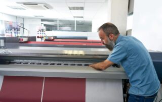 A man stands at a large printout on a wide format printer.