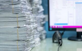 Stacks of paper in the foreground and a blurry computer monitor in the background to signify the need for a document scanner.