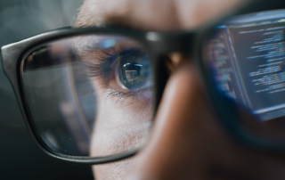 Close-up of a man's upper face. He is wearing glasses and in the reflection of those glasses you can see a computer screen with code to signify cybersecurity.