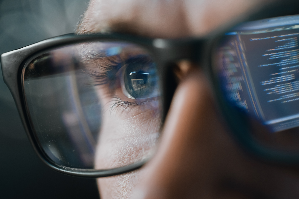 Close-up of a man's upper face. He is wearing glasses and in the reflection of those glasses you can see a computer screen with code to signify cybersecurity.