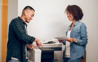 Two diverse employees chat and stand next to an office copier and discuss the interface and outputs.