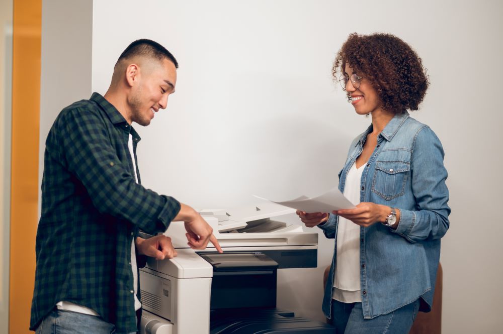 Two diverse employees chat and stand next to an office copier and discuss the interface and outputs.