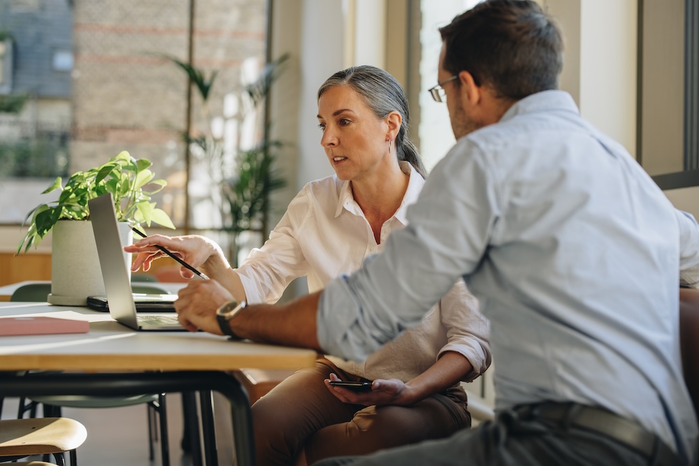 Woman and man working on project at office desk. Coworkers using laptop and discussing work in office.